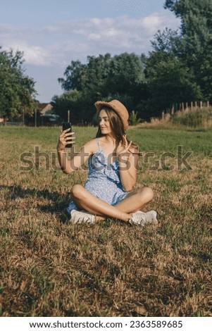 Beautiful caucasian girl with long hair in the blue dress and summer hat filming a video and articulates with her hand at countryside. Vertical photo, social networks concept.