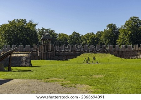Abstract photo of human settlement exposure. Grassy landscape, wooden palisades made of wooden, sharp stakes. Royalty-Free Stock Photo #2363694091
