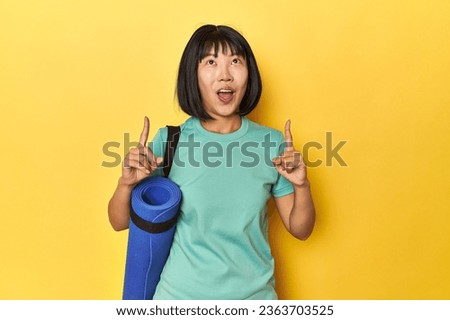 Chinese woman, sportswear and mat, in yellow studio pointing upside with opened mouth.