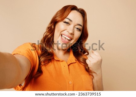Close up happy young ginger chubby overweight woman wears orange shirt casual clothes doing selfie shot pov on mobile cell phone do winner gesture isolated on plain beige background studio portrait