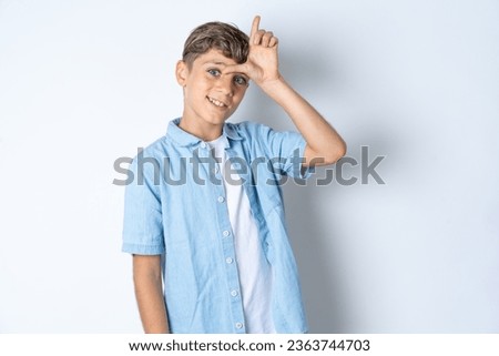 Handsome Caucasian teenager boy wearing denim jacket making fun of people with fingers on forehead doing loser gesture mocking and insulting.