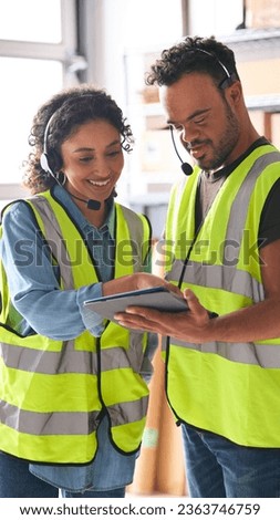 Woman Using Digital Tablet And Male Colleague With Down Syndrome In Warehouse Wearing Headsets Royalty-Free Stock Photo #2363746759