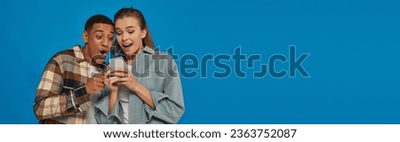amazed interracial couple looking at smartphone on blue backdrop, social media users, banner Royalty-Free Stock Photo #2363752087