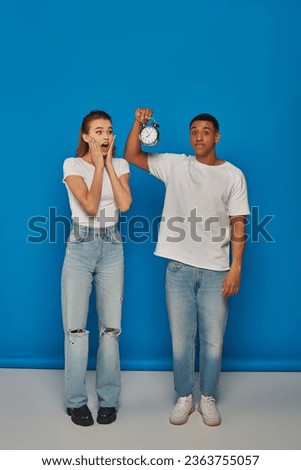 morning time, african american man holding alarm clock near astonished woman on blue backdrop