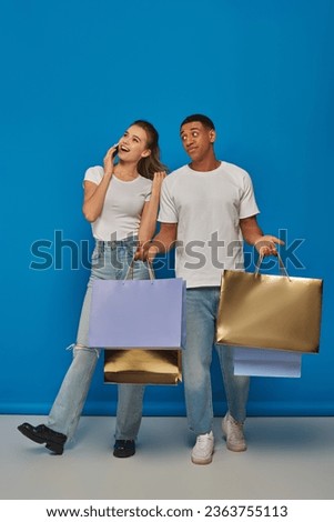 consumerism, interracial couple  holding shopping bags on blue backdrop, woman talking on smartphone