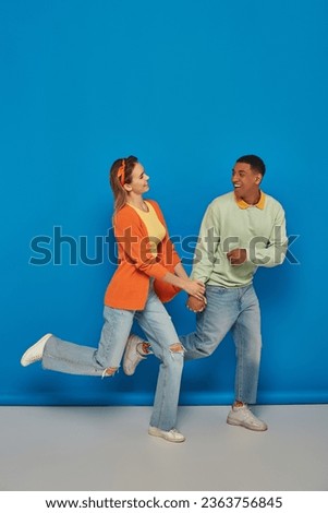 happy multicultural couple in casual attire holding hands and running on blue background, smile
