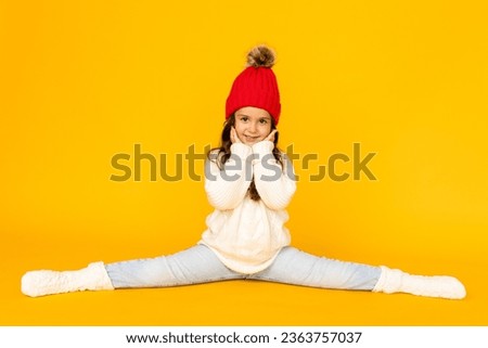 Funny child girl in a white sweatshirt and red knitted hat with pompom doing twine on a yellow background. The concept of children's emotions, joy and delight. Advertising clothing and sales.