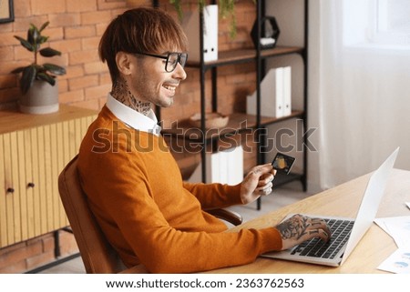 Young man with credit card using laptop at table in office
