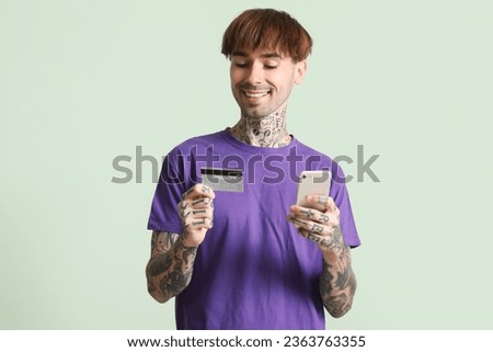 Tattooed young man with credit card using mobile phone on green background
