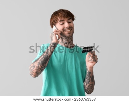 Tattooed young man with credit card talking by mobile phone on light background