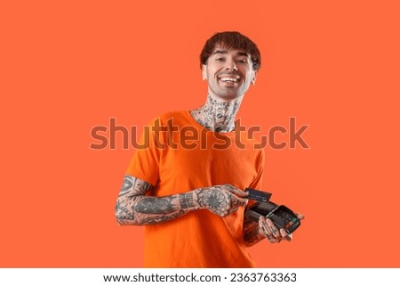 Tattooed young man paying with credit card via terminal on orange background