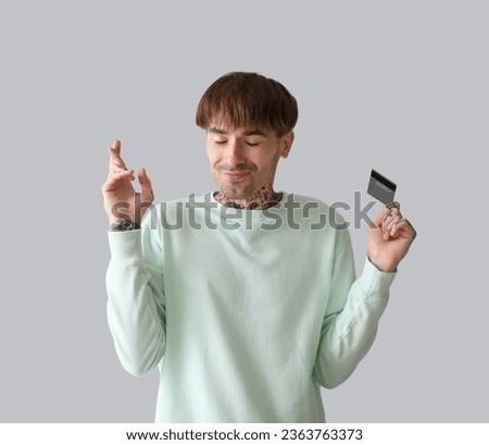 Tattooed young man with credit card crossing fingers on light background