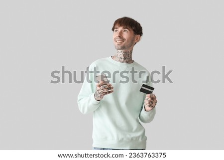 Tattooed young man with credit card and mobile phone on light background