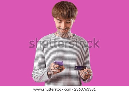Tattooed young man with credit card using mobile phone on purple background