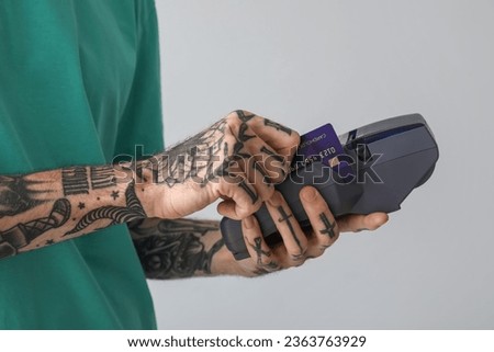 Tattooed young man paying with credit card via terminal on light background, closeup
