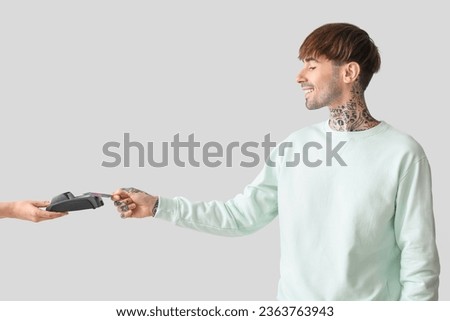 Tattooed young man paying with credit card via terminal on light background
