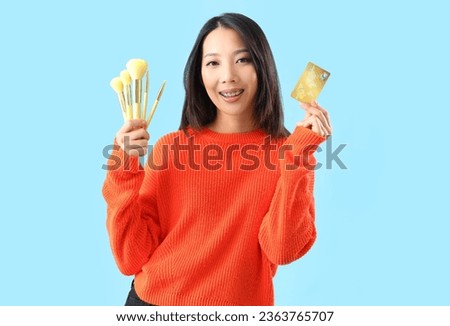 Young Asian woman with credit card and makeup brushes on blue background