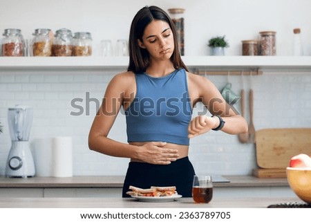 Shot of hungry sporty woman waiting for the time to eat while looking watch after fasting in the kitchen at home Royalty-Free Stock Photo #2363789705