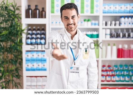 Handsome hispanic man working at pharmacy drugstore smiling cheerful offering palm hand giving assistance and acceptance. 