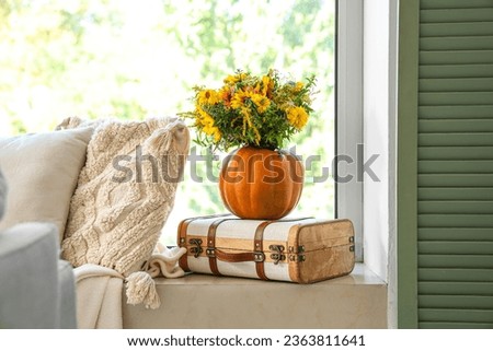Pumpkin with autumn flowers on suitcase near window in room