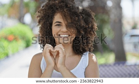 Young beautiful hispanic woman smiling confident doing heart gesture with hands at park