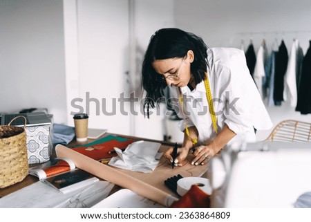 Concentrated young female seamstress in casual wear creating patterns with pencil and measuring tape during work on new design project of clothes. Successful businesswoman designer posing for camera