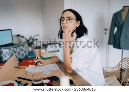 Thoughtful female designer in casual wear and eyeglasses looking away while working on design project at workplace with sewing supplies