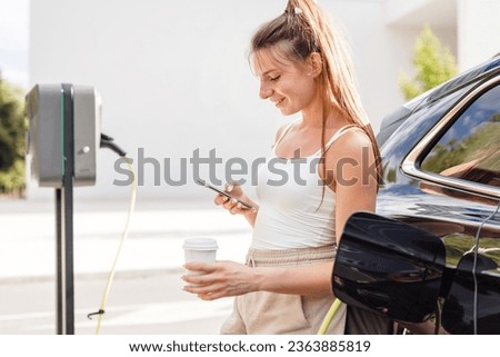 Young woman, putting a charger in an electric car parked in the parking area and adjusting an EV charging app on a mobile phone