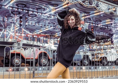 Young cool woman with curly hair and bright make-up in sunglasses having fun with penny board in amusement park