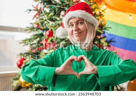 Adult woman makes heart sign with his hands Christmas tree at home. Happy middle-aged female, lgbt concept