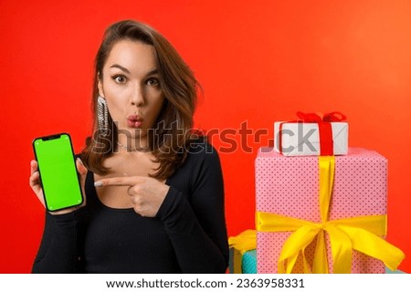 young cute european woman in a black blouse rejoices in gifts holding a smartphone with a mockup on a red background.