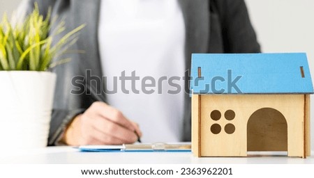 House model on the table, blurred woman signing mortgage loan agreement on background. Concept of home loan and insurance. Buying or renting house, real estate. Selective focus