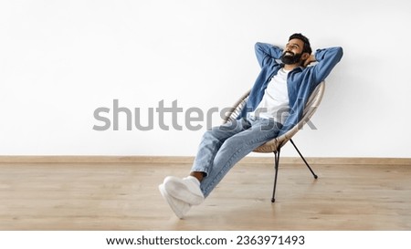 Relaxed young Indian man relaxing with hands behind head in armchair indoor, enjoying comfort and rest on chair near white studio wall. Panorama with empty space for text advertisement