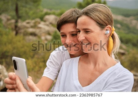 Happy adult mother and teenage son sitting with smartphone in daylight and attending video call