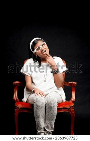 Embodying a book-smart but socially naive stereotype, an Indian woman in white glasses and headband jestingly acts clueless, seated on an aged chair Royalty-Free Stock Photo #2363987725