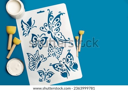 Reusable flexible plastic stencil for graphical design, white paint and round sponge foam brushes on a blue background Royalty-Free Stock Photo #2363999781