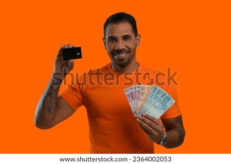 man holds a black credit card and Brazilian money, wearing an orange shirt on an orange background