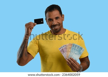 man holds a black credit card and Brazilian money, wearing a yellow shirt on a blue background