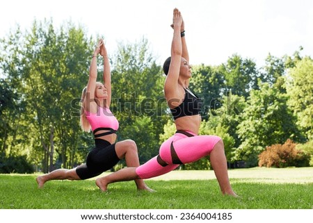 Two young women practice yoga in summer park. Slender caucasian girls in pink-black bodysuits doing exercises to strengthen their body and spirit. Morning gymnastics in fresh air. Healthy lifestyle.