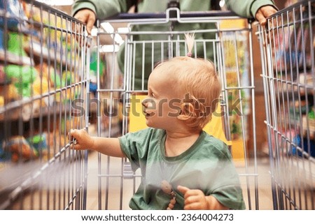 Close-up of cute baby in shopping cart being driven by her mom in supermarket. Mother and little blonde daughter in similar green clothes shopping and having fun. Family weekend, happy childhood.