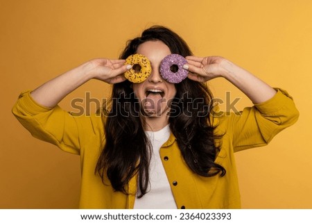 Waist-up portrait of a mischievous lady looking through the holes in the cookies while showing her tongue to the camera. Unhealthy eating concept