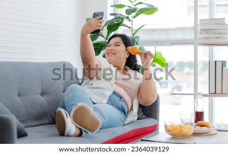Asian young cheerful overweight oversized fat chubby plump unhealthy female teenager in casual outfit laying lying down smiling on cozy sofa holding croissant taking selfie photo via smartphone.