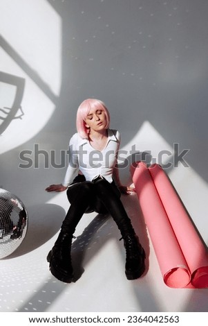 A young woman with pink hair sits elegantly in a minimalist studio, wearing a white blouse, black pants, and leather boots. Modern style and individuality, suitable for fashion and editorial concepts