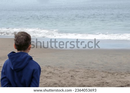 Portrait of a 10-year-old blond boy on the sandy beach on a cloudy day. The boy is dressed in a warm sweatshirt. He sits with his back to us. Royalty-Free Stock Photo #2364053693