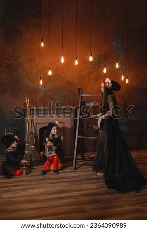 Girls in witch costumes are sitting near the fireplace on pumpkins, one is holding a lantern, the other is making a potion, and their mother is standing nearby. Halloween. Vertical frame.