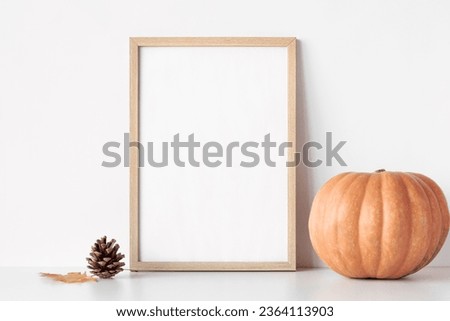 Empty wooden frame stands on white table with pumpkin, pinecone and yellow leaf. Autumn mockup poster frame close up.