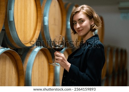 Professional Caucasian woman sommelier tasting and smelling red wine in wine glass at wine cellar with wooden barrel at wine factory. Alcohol liquor shop, winery industry and winemaker concept. Royalty-Free Stock Photo #2364159893