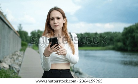 A beautiful young teenage girl in a white top, with long brown hair, uses a smartphone outdoors in the summer, while walking in nature on the shore of a lake. The girl writes text messages and