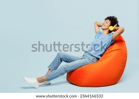 Full body young woman of African American ethnicity wear t-shirt casual clothes sit in bag chair listen to music in headphones isolated on plain pastel light blue cyan background. Lifestyle concept
