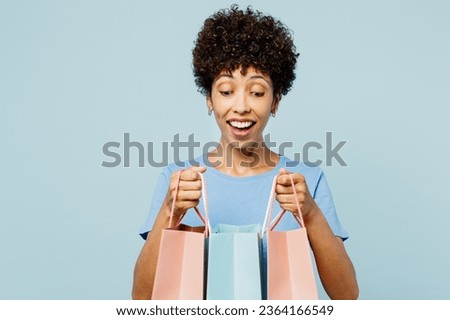 Young shocked woman of African American ethnicity wearing casual clothes hold in hand paper package bags after shopping isolated on plain light blue cyan background. Black Friday sale buy day concept